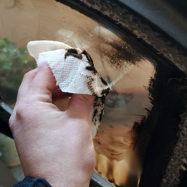 hand holding wet paper towel, wiping the inside glass of a pellet stove. glass is coated with black carbon deposits from fire.