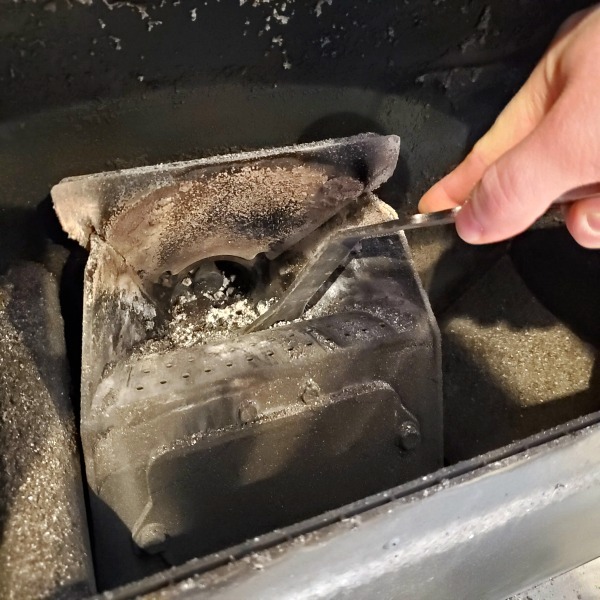 scraping the inside of a Harman pellet stove burn pot with a scraper. Ash can be seen inside the neck of the burn pot near the auger.